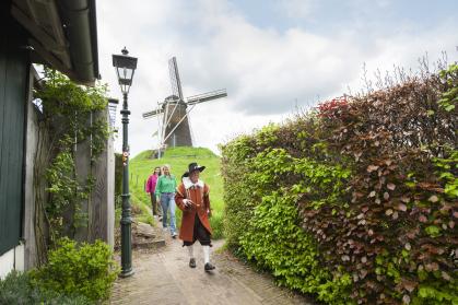 Wandelaars lopen met een gids in historische kleding door een smal straatje in Bredevoort, met op de achtergrond een molen op een groene heuvel.
