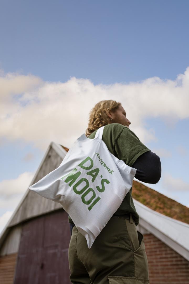 Een man staat voor een boerderij en draagt een tas met de tekst 'Da's mooi'