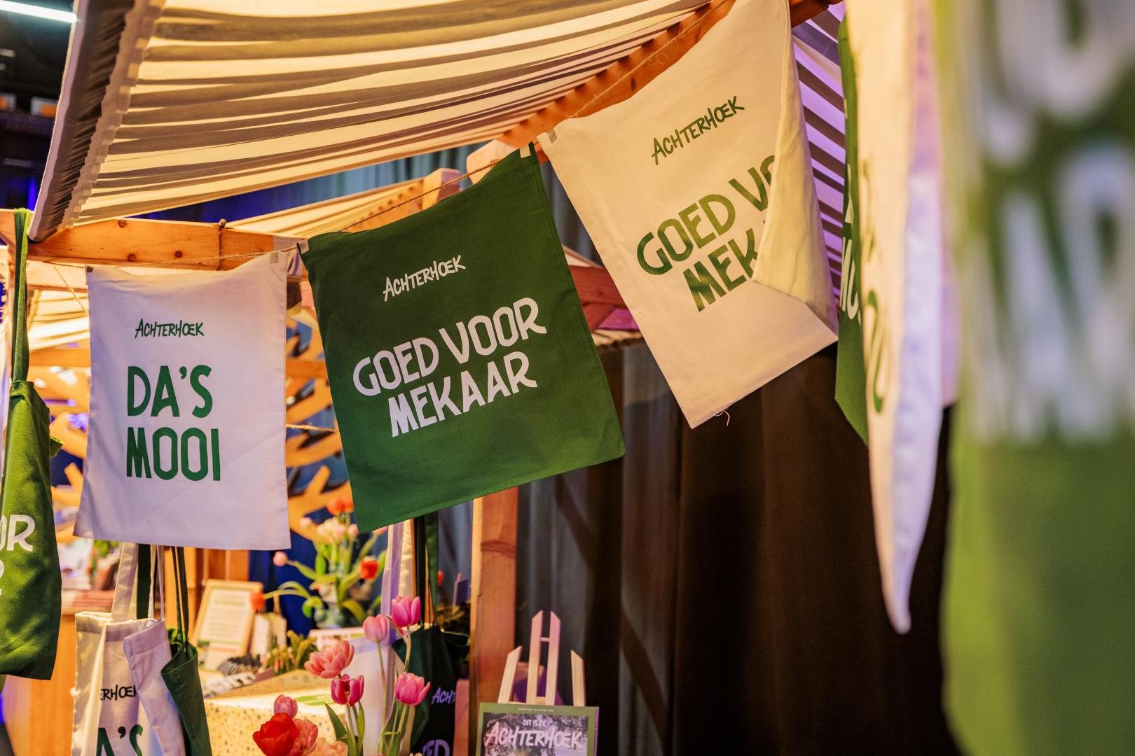 Aan een marktkraam hangen tasjes van Achterhoek Toerisme met de teksten 'Goed voor mekaar' en 'Da's mooi'.