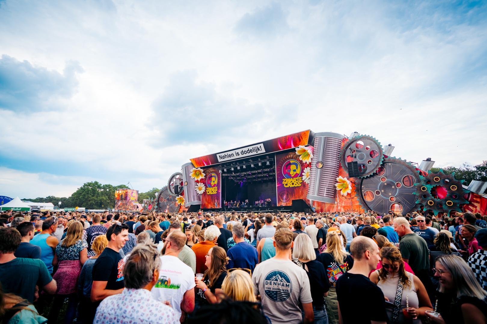 Bezoekers staan voor het hoofdpodium van de Zwarte Cross tijdens een optreden. Voor het grote, kleurrijke podium heeft zich een grote menigte verzameld onder een bewolkte lucht.